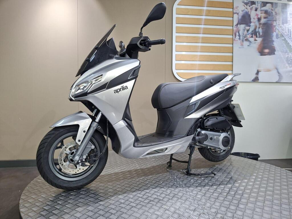 Aprilia SXR 50 2026 8 A silver Aprilia scooter showcased on a textured display platform, set against a light-colored wall with a blurred background image of people walking.