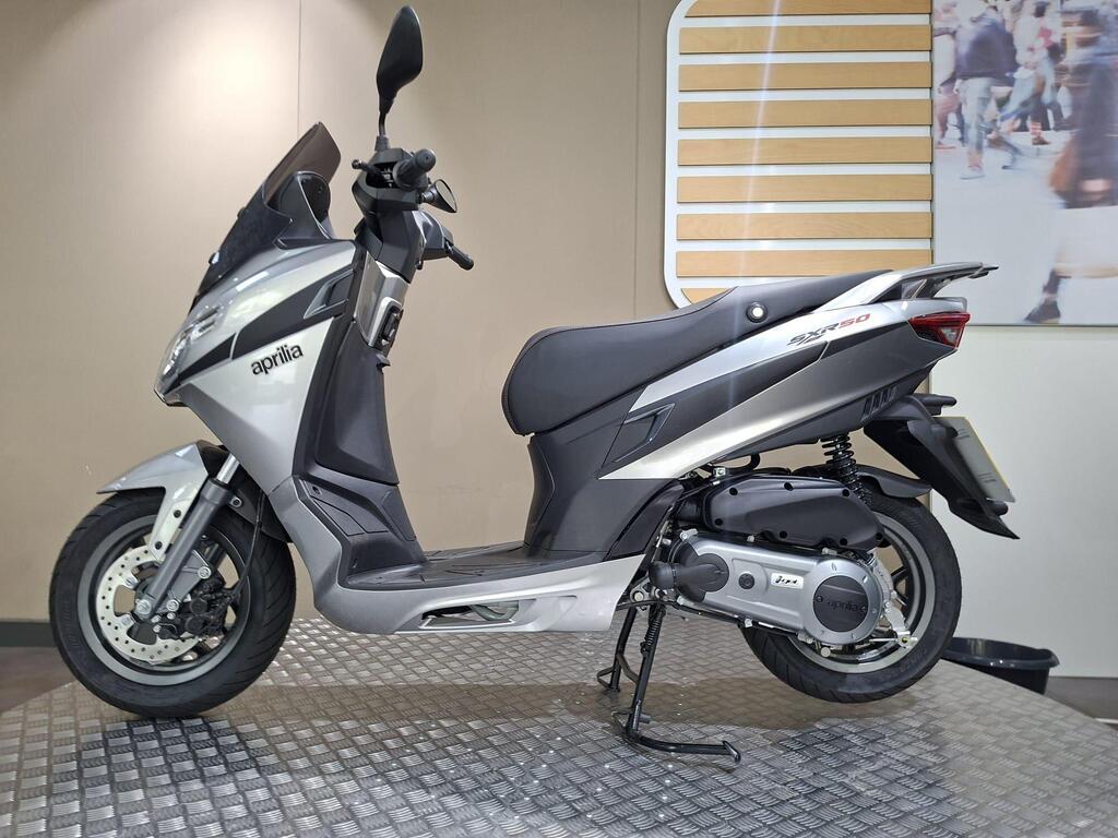 Aprilia SXR 50 2026 9 A silver Aprilia SR 50 scooter positioned on a textured floor with a minimalist background featuring a light-colored wall and a graphic design element.