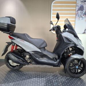 Piaggio MP3 300 Sport 2021 A sleek grey three-wheeled scooter with a large rear storage box, displayed on a textured black surface, set against a neutral wall with a blurred urban scene in the background.