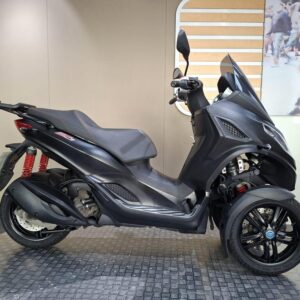 Piaggio MP3 300 Sport 2022 A black three-wheeled scooter is displayed on a textured surface, showcasing its sleek design and red shock absorbers. The background features a simple, neutral wall that enhances the scooter's profile.