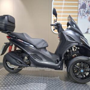 Piaggio MP3 300 Sport 2020 A black three-wheeled scooter is displayed on a textured circular platform, featuring a sleek design, a rear storage box, and red suspension springs. The background includes a softly lit wall with a modern pattern and a blurred image of people.
