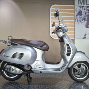 The Vespa GTS 300 Super 2016 A silver Vespa scooter with a brown seat is displayed against a neutral background. The scooter features a classic design with a chrome rearview mirror and distinctive body lines.