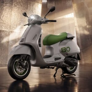 Vespa Edizione Ottantesimo A modern scooter with a sleek silver body and a green seat, set against a glossy, metallic background, showcasing its stylish design.
