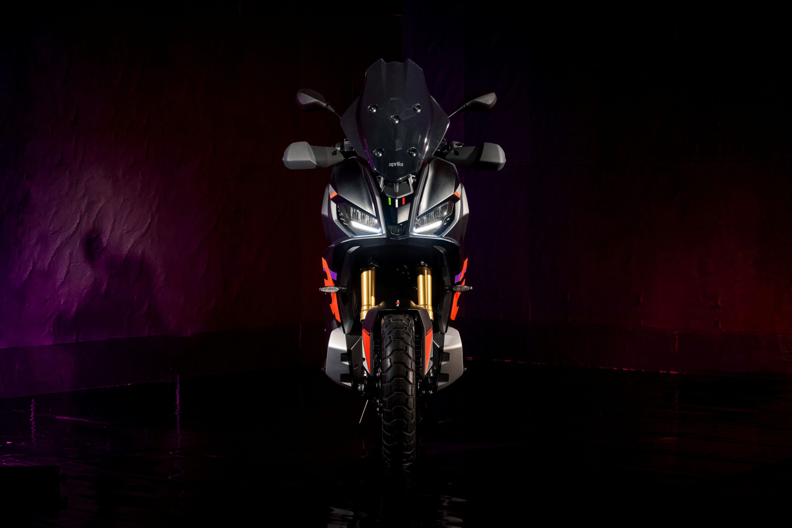Aprilia SRGT400 Rally Replica Front A sleek modern motorcycle with a distinctive design, featuring a matte black body, prominent front headlights, and striking orange accents, set against a dark background with colorful lighting.