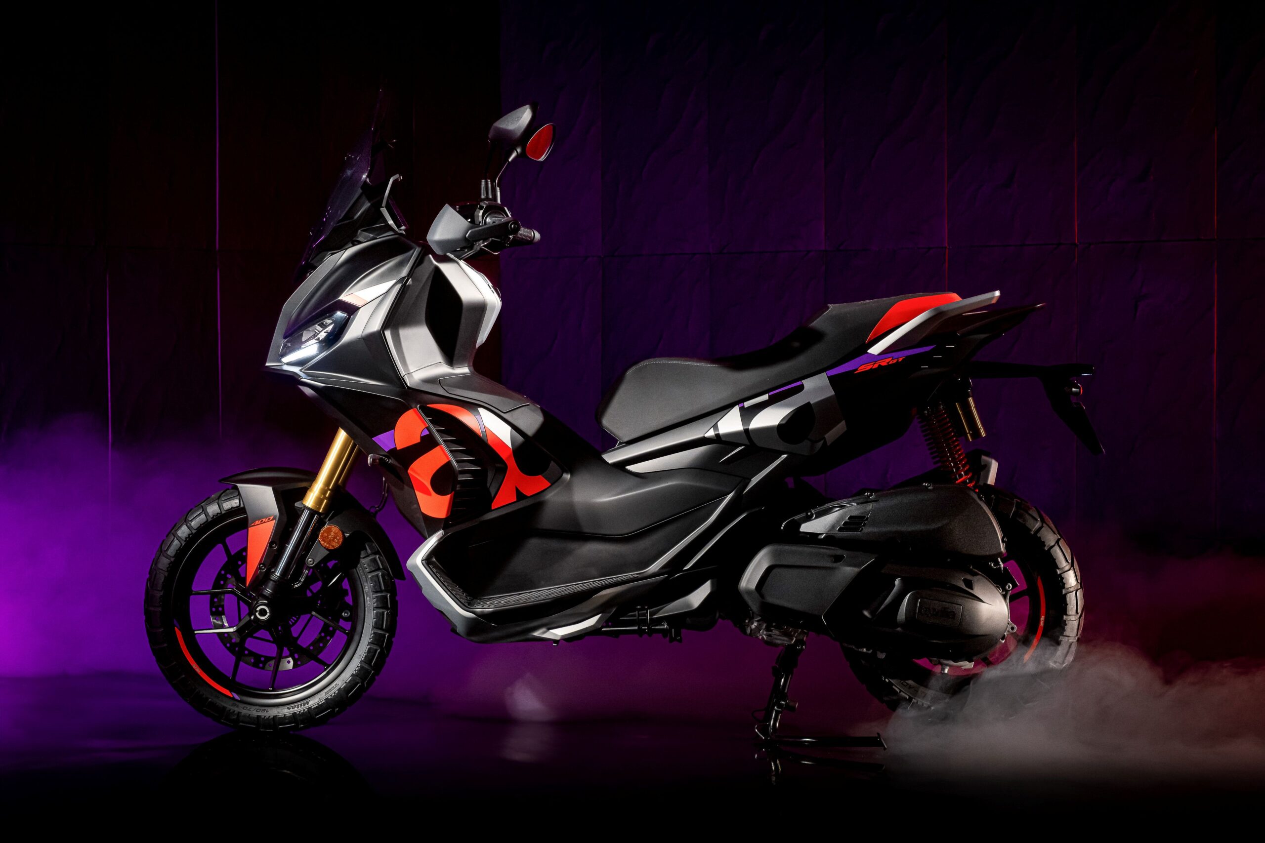 Aprilia SRGT400 Rally Replica Left A sleek, modern motorcycle with a black and red design, showcasing a prominent front headlight and a sporty silhouette against a moody purple background with smoke effects.