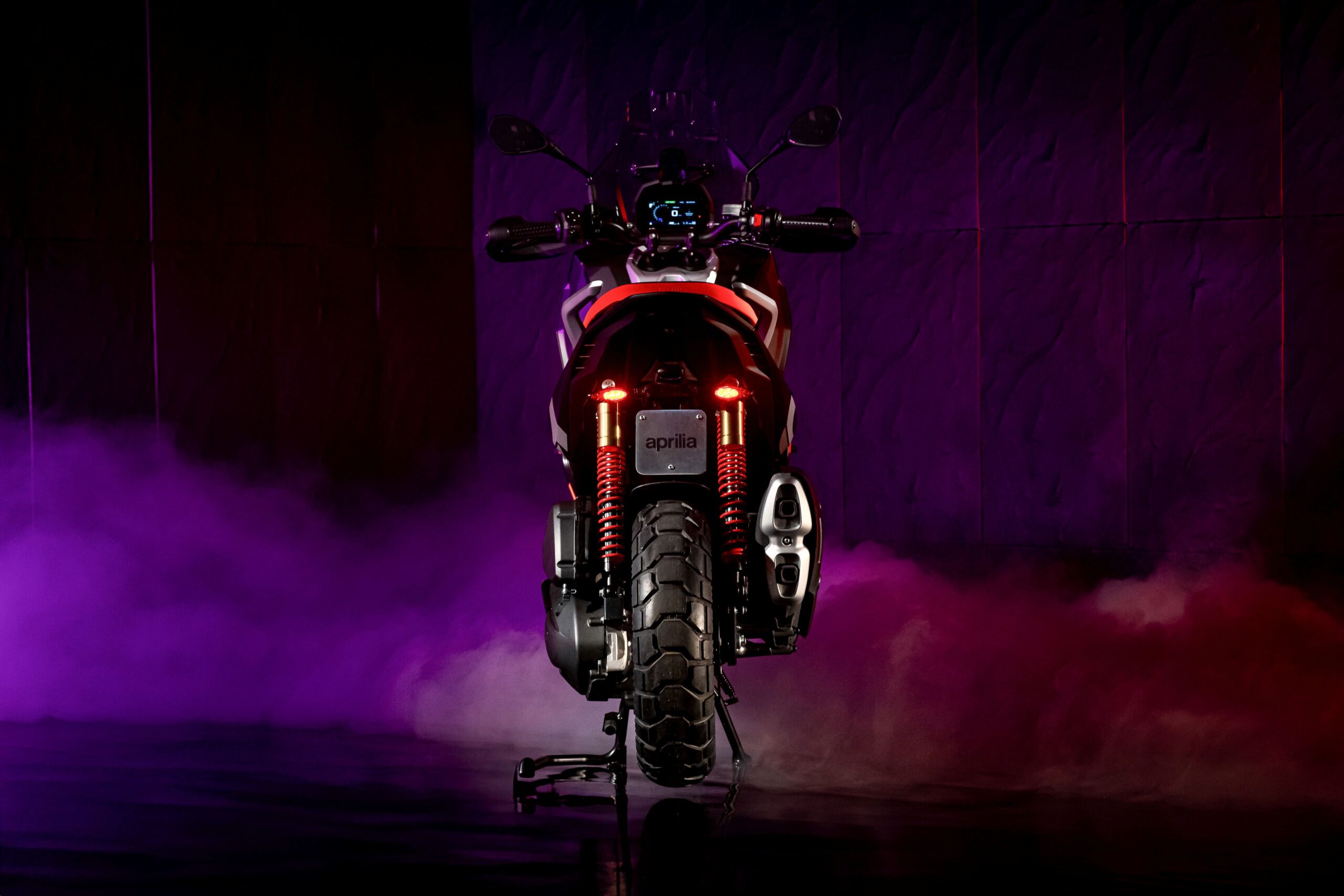 Aprilia SRGT400 Rally Replica Rear A rear view of a motorcycle, illuminated with purple and pink lighting, showcasing its sleek design, including a prominent tail with red lights and a patterned rear tire. Smoke adds a dramatic effect to the scene.