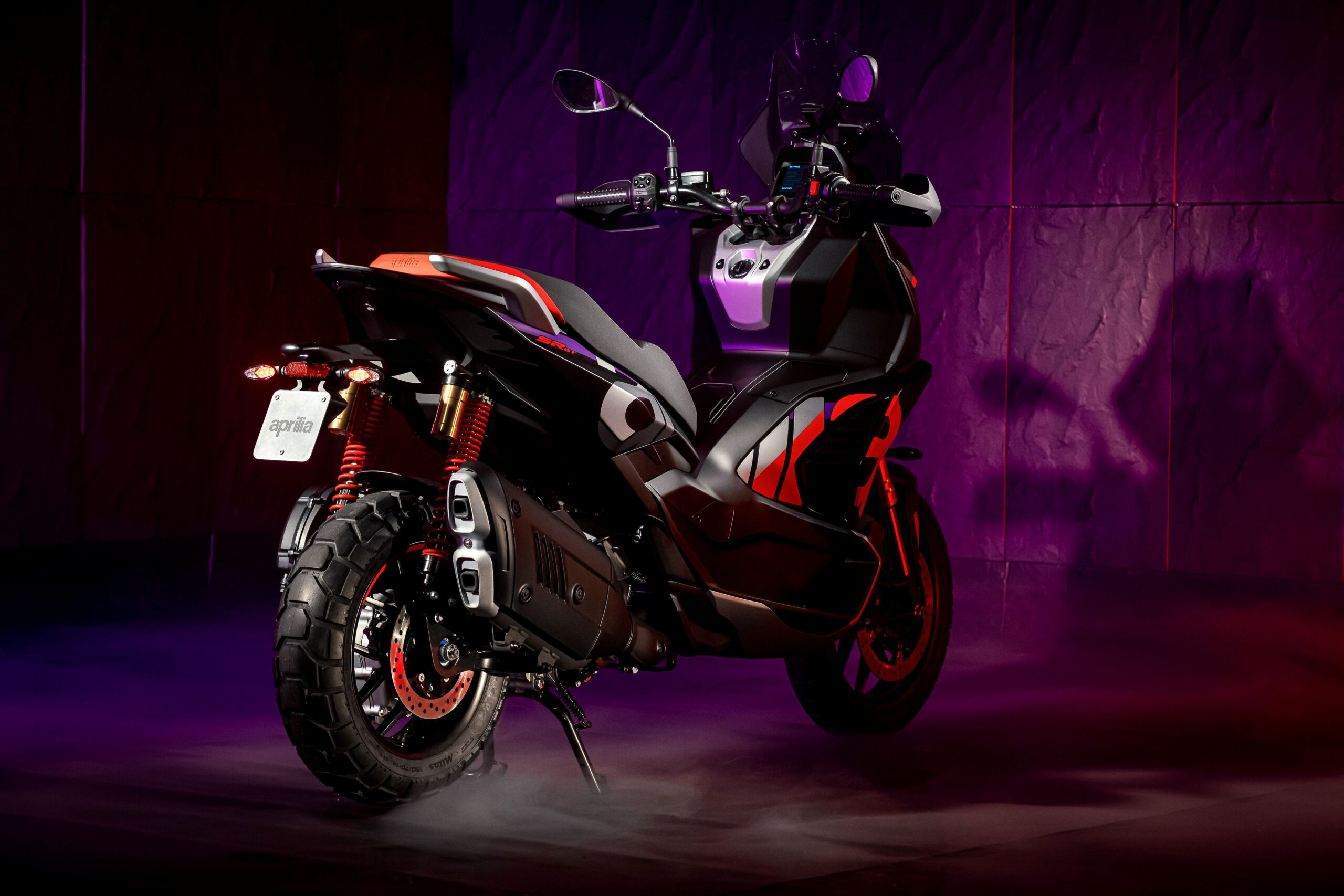 Aprilia SRGT400 Rally Replica Right Rear A sleek black and red scooter displayed against a gradient purple background, showcasing its modern design and details such as pronounced tires and dual exhaust pipes.