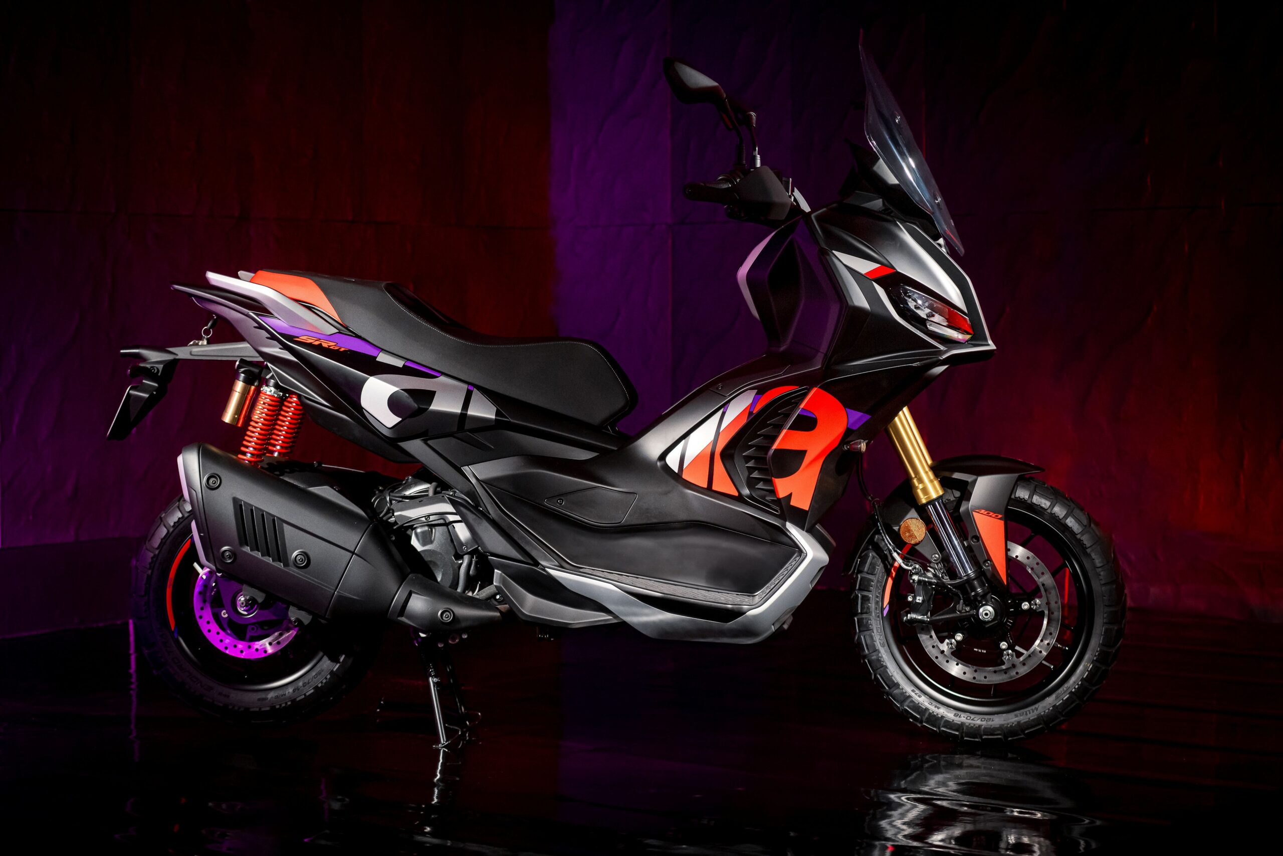Aprilia SRGT400 Rally Replica Right Aprilia SR GT 400 Rally Replica A sleek, modern scooter in black and orange, featuring a sporty design with a prominent front headlight, a high windshield, and distinctive color accents, set against a dramatic purple and red backdrop.
