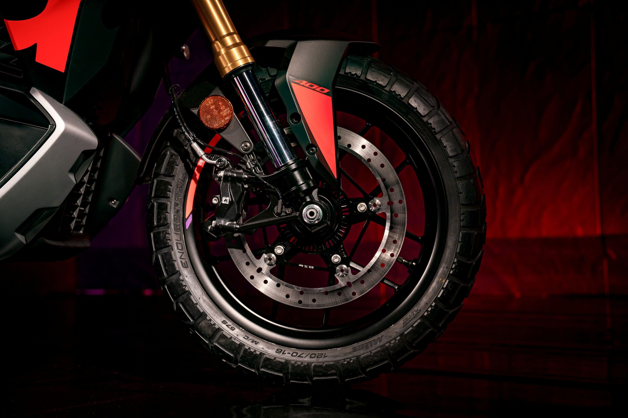 Aprilia SRGT400 Rally Replica Wheel Close-up of a motorcycle's front wheel, featuring a detailed tread pattern, disc brake, and suspension elements, set against a gradient background with red and purple hues.