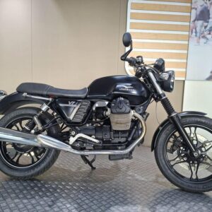 A black Moto Guzzi V7 motorcycle is displayed on a textured surface with a beige wall in the background. The bike features a retro design with chrome accents, a low saddle, and a prominent engine.