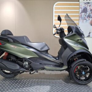 A green three-wheeled scooter displayed on a textured platform inside a showroom, featuring a streamlined design, sleek seat, and prominent rear suspension.