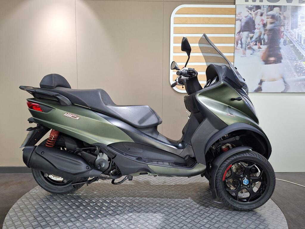 Piaggio MP3 350 LT Sport 2020 1 A green three-wheeled scooter displayed on a textured platform inside a showroom, featuring a streamlined design, sleek seat, and prominent rear suspension.