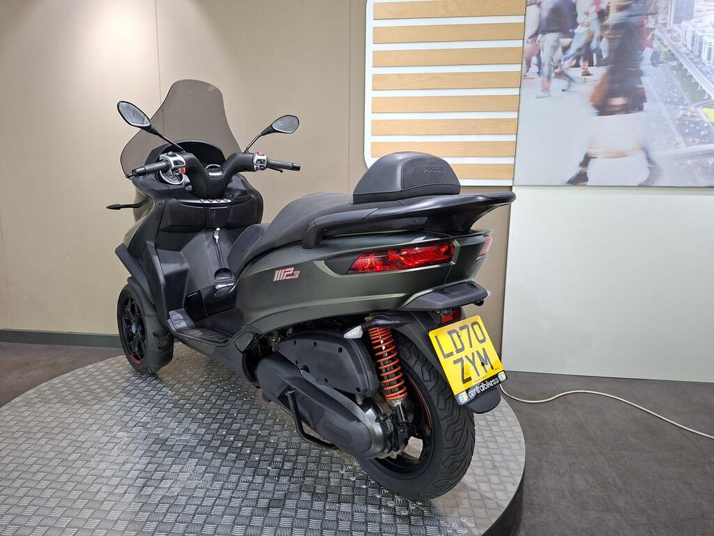 Piaggio MP3 350 LT Sport 2020 10 A three-wheeled scooter displayed in a showroom, featuring a sleek dark design, windshield, and a visible license plate reading "LD70 ZYM." The background includes a wall with wooden accents and a blurred image of people walking.