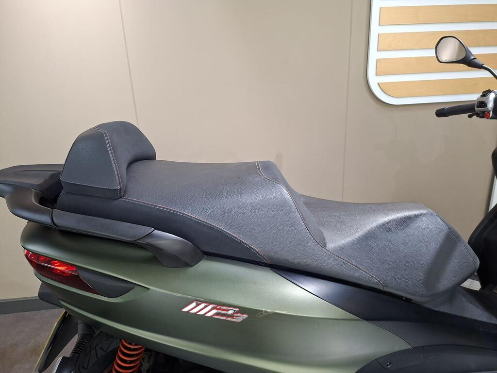Piaggio MP3 350 LT Sport 2020 12 A close-up view of a motorcycle seat, featuring a black textured surface with red stitching, mounted on a green scooter body with visible suspension components. The background is a neutral-colored wall with wooden accents.