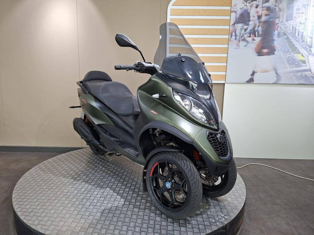 Piaggio MP3 350 LT Sport 2020 2 A green three-wheeled scooter with a sleek design is positioned on a circular platform with a metallic surface. The scooter features a windshield, sporty black wheels, and a comfortable seat. In the background, there is a display of people walking in an urban setting.