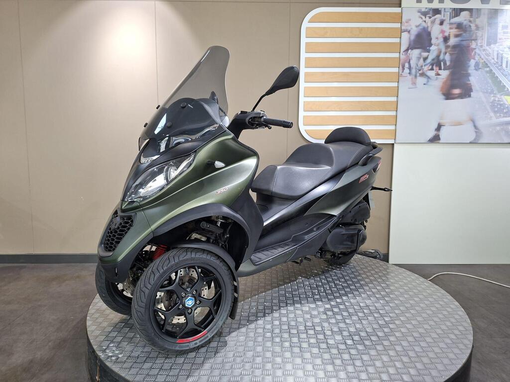 Piaggio MP3 350 LT Sport 2020 4 A green three-wheeled scooter displayed on a circular platform, featuring a front windshield, black seat, and alloy wheels, with a modern background showing a cityscape.