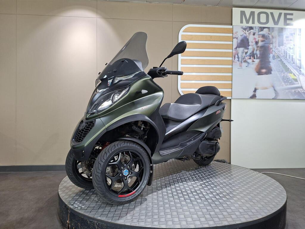 Piaggio MP3 350 LT Sport 2020 5 A green three-wheeled scooter displayed on a circular platform with a textured surface, featuring a transparent windshield, sleek design, and sporty black wheels. The background includes promotional signage and a blurred image of people walking.
