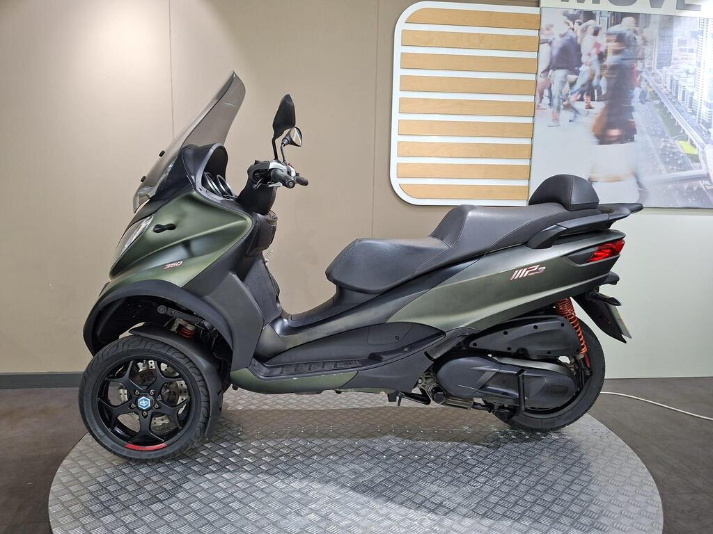 Piaggio MP3 350 LT Sport 2020 6 A dark green three-wheeled scooter displayed on a circular platform, featuring a sleek design, black wheels, and a transparent windshield, set against a neutral background with a modern wall graphic.