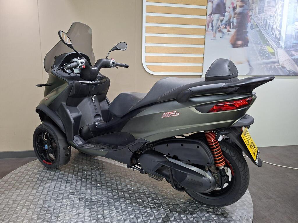 Piaggio MP3 350 LT Sport 2020 7 A three-wheeled scooter with a modern design, featuring a dark gray body, a tinted windshield, and sporty accents, positioned on a textured circular display platform. In the background, there is a blurred image of a cityscape with people walking.
