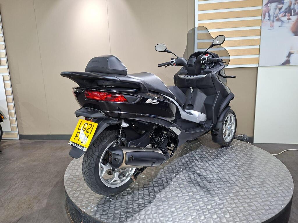 Piaggio MP3 500 Business 2021 10 A black three-wheeled scooter is displayed on a circular stand in a showroom, showcasing its rear and side profile; a smooth floor and neutral-toned walls are in the background.