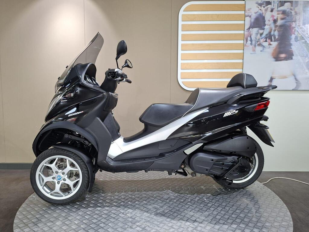 Piaggio MP3 500 Business 2021 11 A black three-wheeled scooter displayed on a circular platform with a textured surface, featuring a large windscreen and a sleek design, set against a neutral wall background.