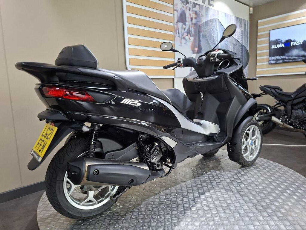 Piaggio MP3 500 Business 2021 12 A sleek black three-wheeled scooter displayed in a showroom, featuring modern design elements and a rear view showcasing its rear tire, exhaust, and seat. The background includes a light-colored wall with promotional graphics.