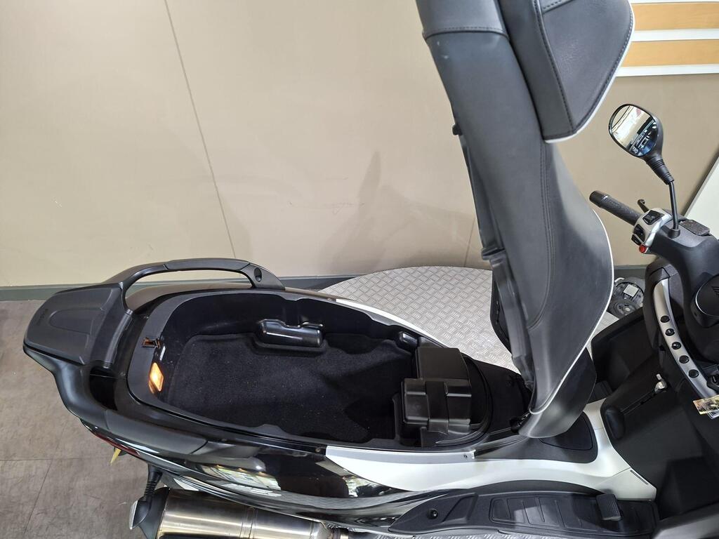 Piaggio MP3 500 Business 2021 14 A top-down view of a scooter's seat with the seat raised, revealing a storage compartment beneath, along with a small black box inside the compartment and controls on the right. The background features a smooth surface and a plain wall.