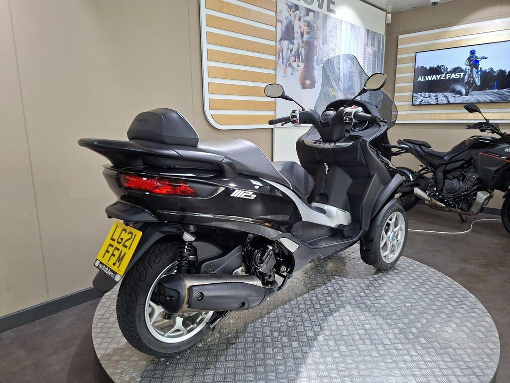 Piaggio MP3 500 Business 2021 15 A black scooter with a windshield, parked on a round metal platform, showcasing its rear. The scooter has a license plate visible with the registration number LG21 FFM, and a light gray wall behind it displaying promotional images of motorcycles.