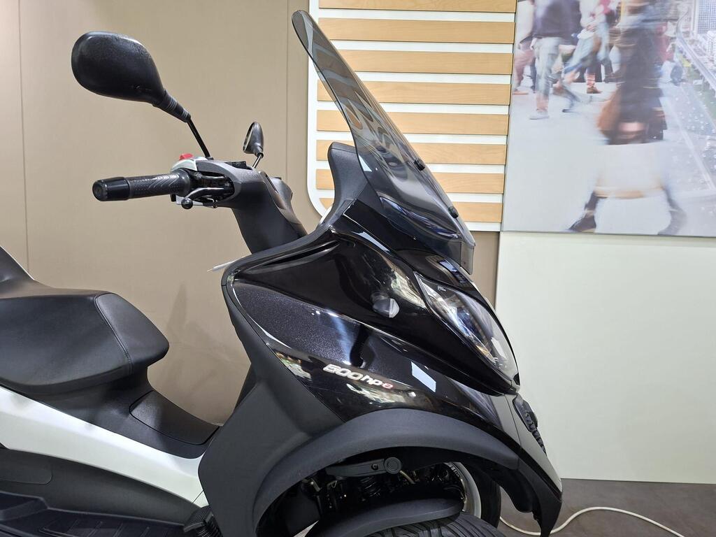 Piaggio MP3 500 Business 2021 16 A close-up view of a black scooter with a windshield, showing the handlebar, mirror, and seat, set against a neutral backdrop featuring a blurred image of people walking.