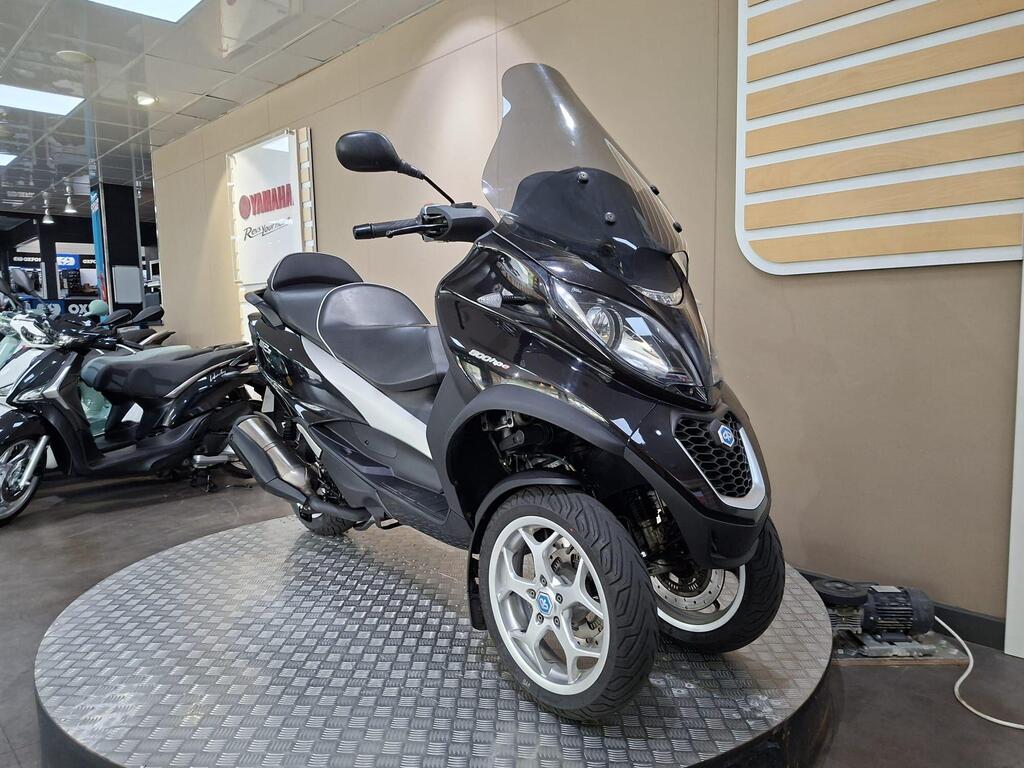 Piaggio MP3 500 Business 2021 17 A black three-wheeled scooter displayed on a circular platform in a showroom, with a transparent windscreen, modern design, and silver rims. Other scooters are visible in the background, along with a Yamaha sign on the wall.