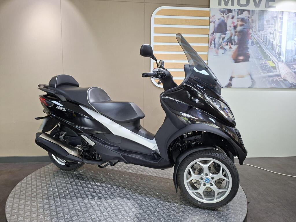 Piaggio MP3 500 Business 2021 2 A black and silver three-wheeled scooter displayed on a circular platform, with a modern backdrop featuring an urban scene.