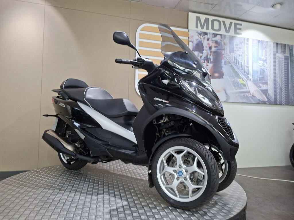 Piaggio MP3 500 Business 2021 3 A black and white three-wheeled scooter displayed on a circular platform, with a modern backdrop featuring the word "MOVE" and a cityscape. The scooter has a streamlined design and large alloy wheels.