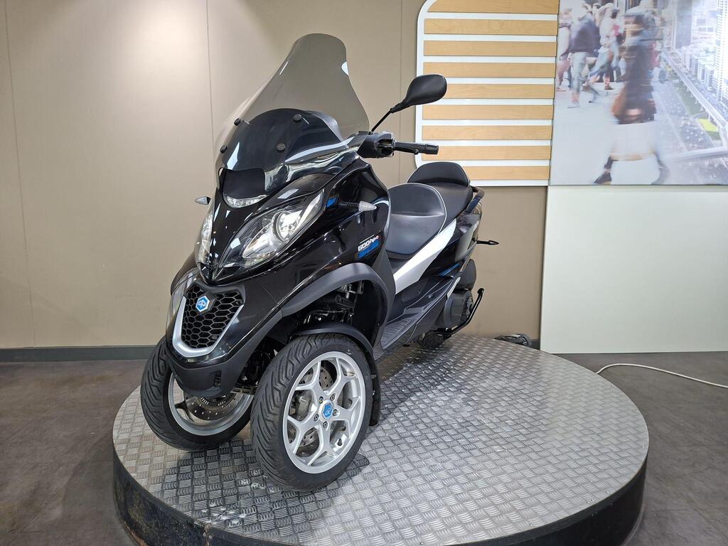 Piaggio MP3 500 Business 2021 7 A black three-wheeled scooter on a circular display platform, featuring a windscreen and modern design elements. The background includes a light-colored wall and a blurred image of pedestrians.