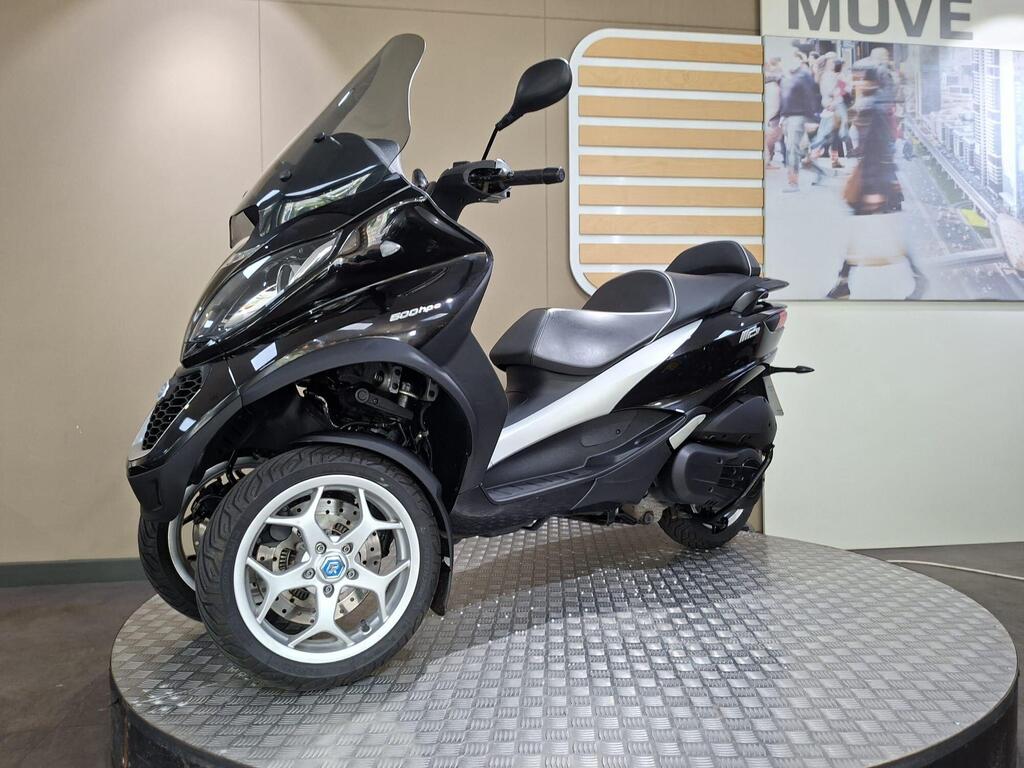 Piaggio MP3 500 Business 2021 8 A black three-wheeled scooter displayed on a round platform with a textured surface, featuring a clear windscreen and a sleek design. The background includes a light-colored wall and a graphic with people walking.