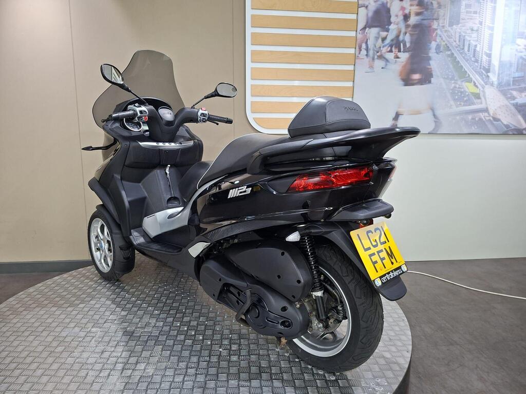 Piaggio MP3 500 Business 2021 9 A black three-wheeled scooter with a windscreen, displayed in a modern showroom with a patterned floor and a partially visible background featuring city pedestrians and buildings. The vehicle has a UK license plate reading "LG21 FFM."