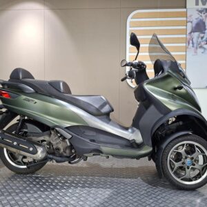 Piaggio MP3 500 LT Sport 2018 A green three-wheeled scooter positioned on a textured platform, featuring a sleek design with a double seat, a windshield, and alloy wheels. The background is a neutral wall with a faint display of pedestrians.