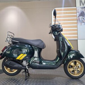 A green vintage scooter with gold wheels, a chrome rear rack, and yellow accents, displayed on a textured surface against a neutral background.