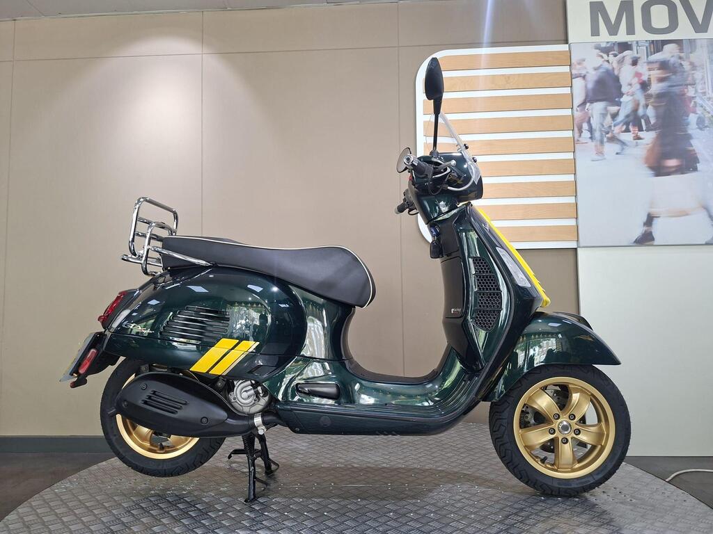 Vespa GTS 300 Racing Sixties 2022 1 A green vintage scooter with gold wheels, a chrome rear rack, and yellow accents, displayed on a textured surface against a neutral background.