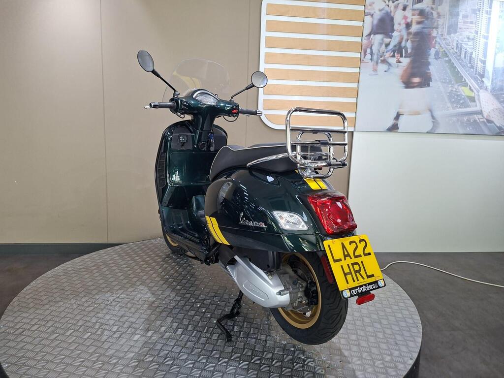 Vespa GTS 300 Racing Sixties 2022 10 A green Vespa scooter displayed on a circular platform, viewed from the rear, featuring a chrome luggage rack and a visible license plate reading "LA22 HRL." The background includes a neutral-colored wall.