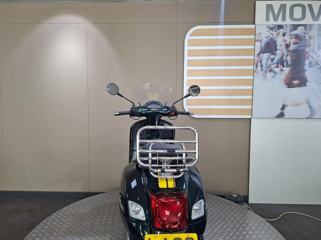 Vespa GTS 300 Racing Sixties 2022 11 A black scooter is showcased from the rear, featuring a chrome rear rack, a clear windshield, and handlebars with mirrors. The background consists of neutral-colored walls and a large photo of a bustling street scene.