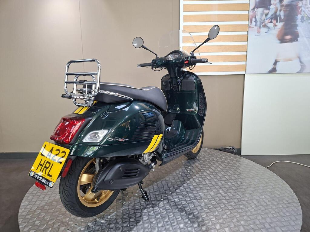 Vespa GTS 300 Racing Sixties 2022 12 A green scooter with gold accents is displayed on a circular platform, featuring a luggage rack and a partial view of a blurred background with people.