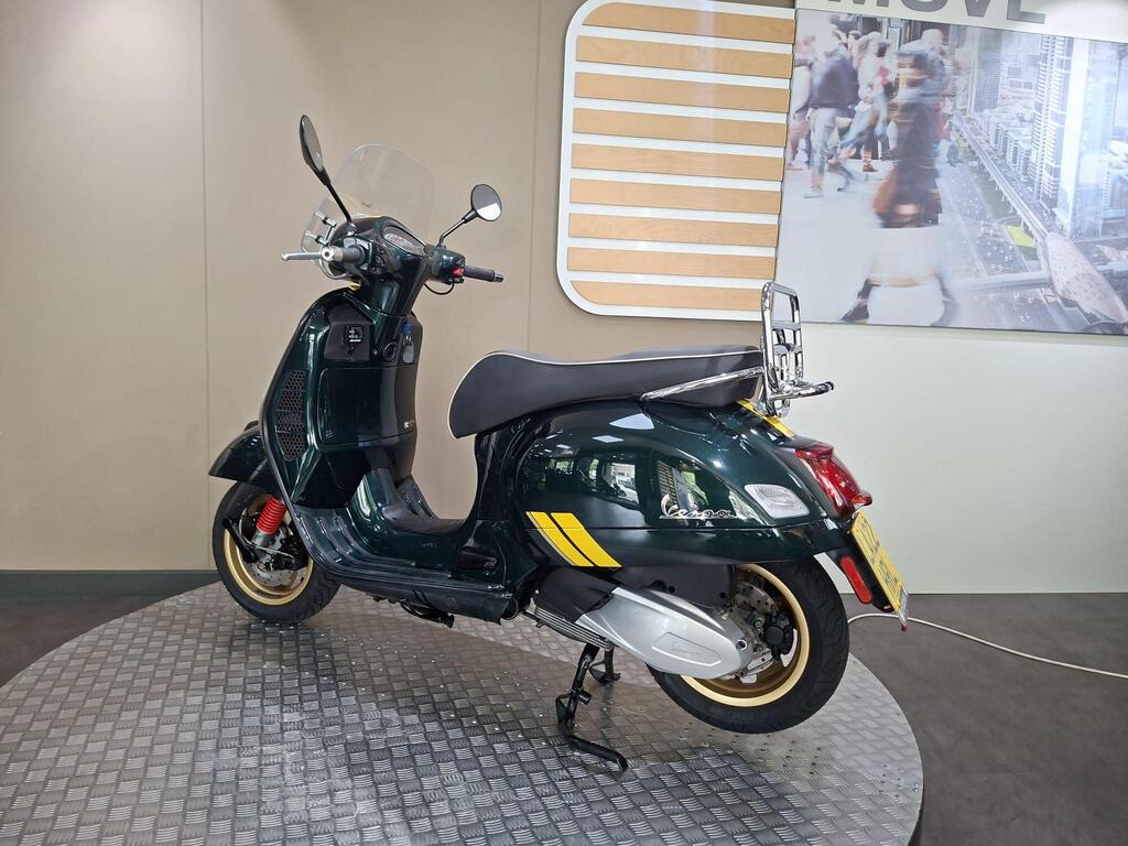 Vespa GTS 300 Racing Sixties 2022 13 A green vintage-style scooter with a windshield and chrome accents is displayed on a circular metallic platform. The background features a wall with the word "MOVE" and an urban scene image.