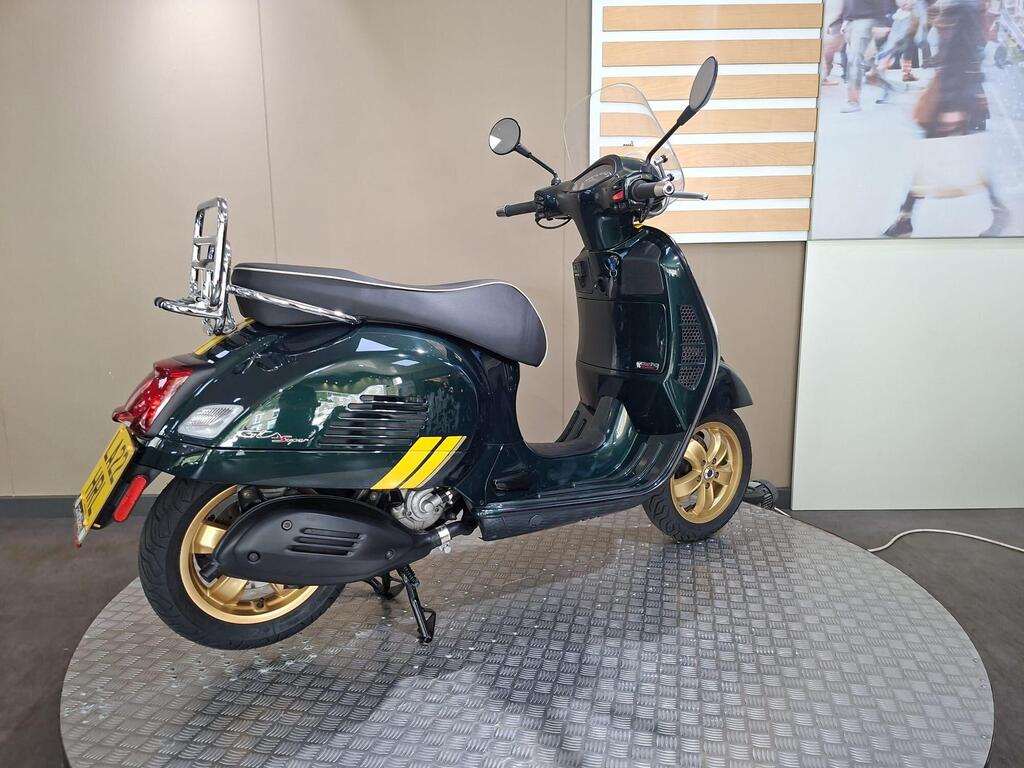 Vespa GTS 300 Racing Sixties 2022 15 A stylish green scooter with gold wheels displayed on a circular platform in a showroom, featuring a chrome rear rack and a transparent windscreen.