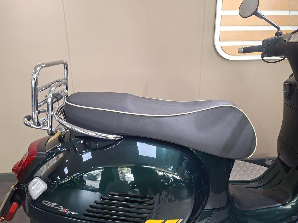 Vespa GTS 300 Racing Sixties 2022 16 A close-up of a green scooter's rear section, featuring a sleek black seat with white piping and a chrome luggage rack attached behind it. The background is a plain light-colored wall.