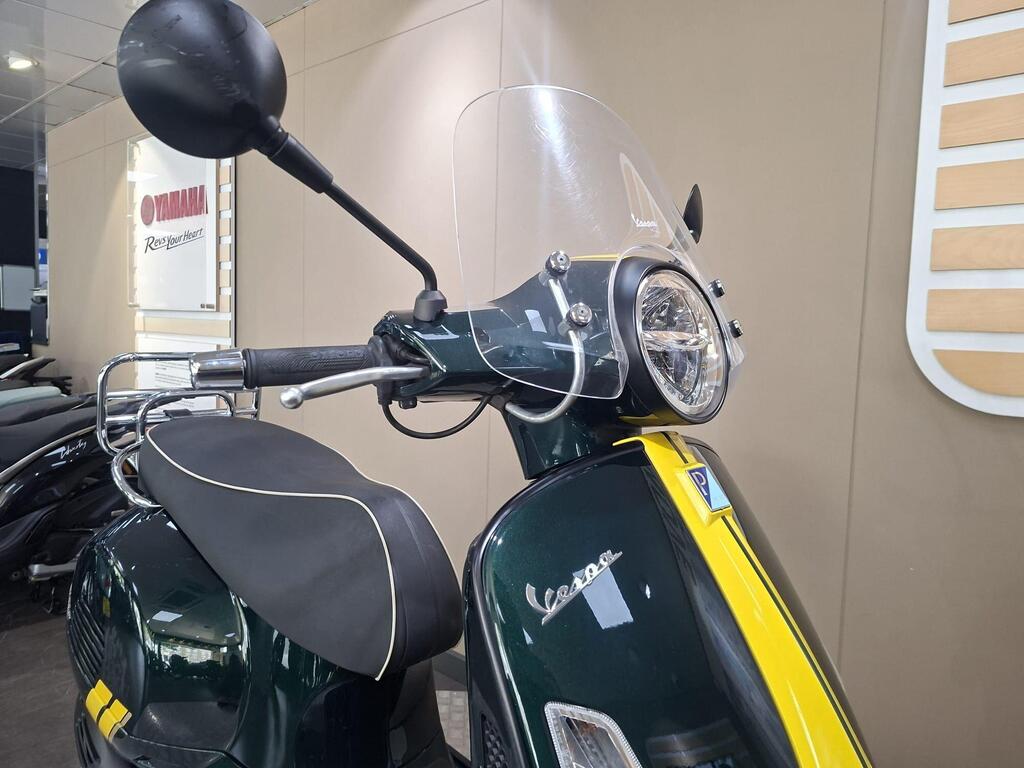 Vespa GTS 300 Racing Sixties 2022 17 A close-up view of a dark green Vespa scooter with a yellow stripe, featuring a transparent windscreen, handlebar with mirrors, and a front headlight, displayed against a neutral background in a showroom.