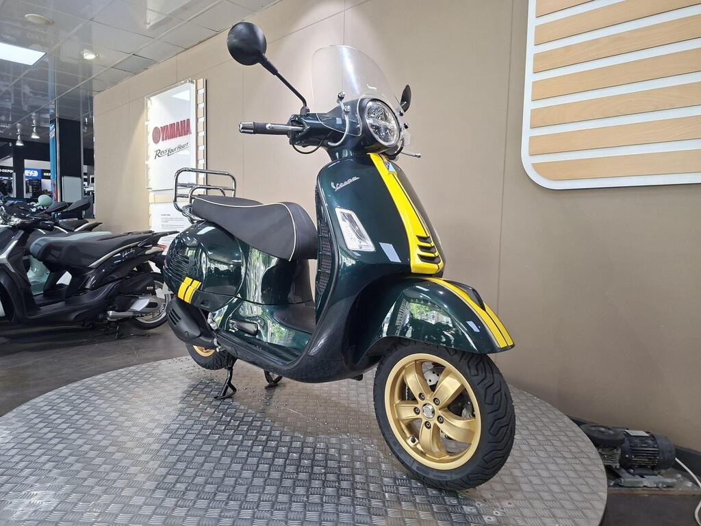 Vespa GTS 300 Racing Sixties 2022 18 A green and yellow Vespa scooter displayed on a circular metal platform in a showroom, with other scooters visible in the background.