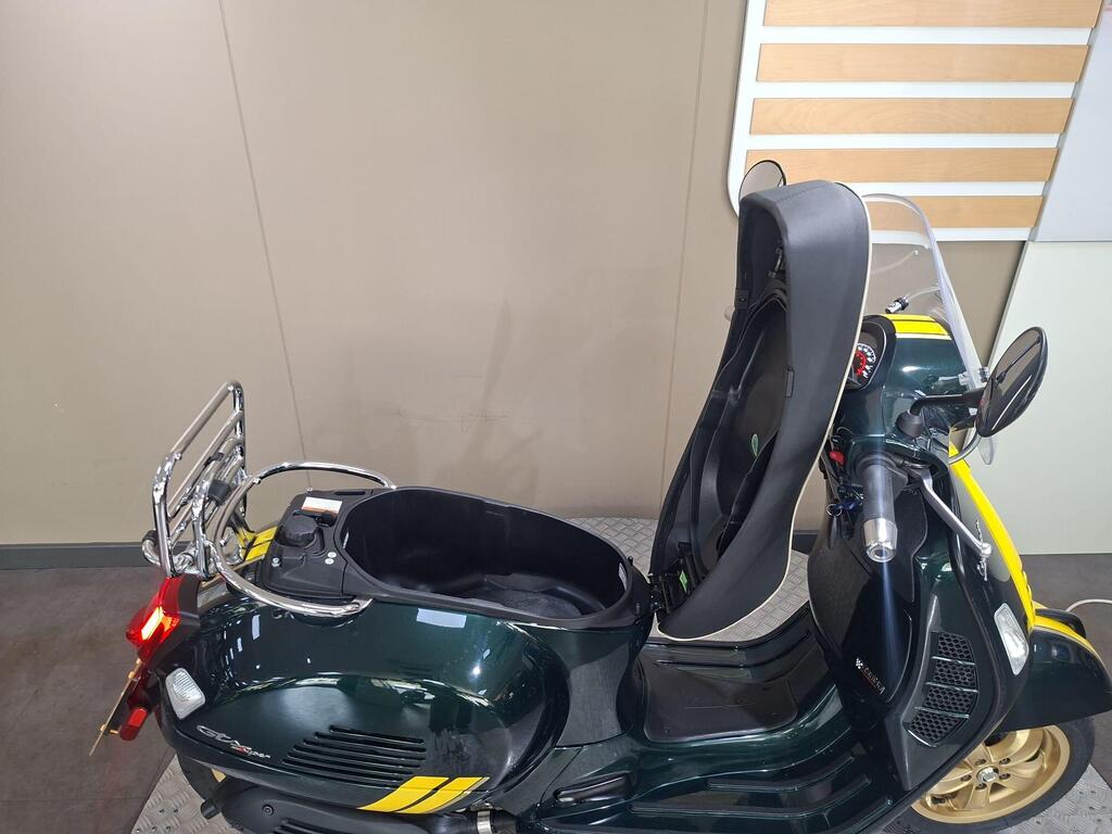 Vespa GTS 300 Racing Sixties 2022 19 A green scooter with yellow stripes featuring a sidecar, displayed in a showroom with neutral-colored walls. The sidecar is empty and visibly spacious.