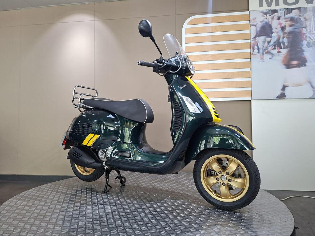 Vespa GTS 300 Racing Sixties 2022 2 A green and yellow Vespa scooter on a circular display stand, positioned against a neutral background with soft lighting. The scooter features gold alloy wheels and a rear luggage rack.