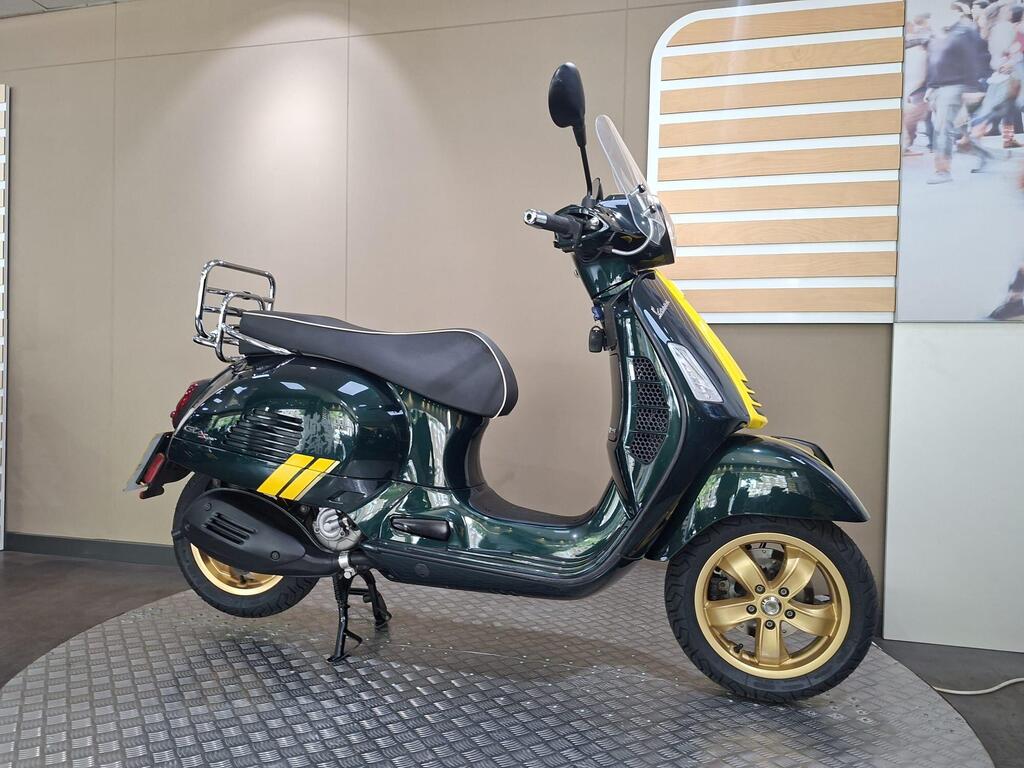 Vespa GTS 300 Racing Sixties 2022 20 A green scooter with gold wheels and a yellow accent, displayed on a circular platform in a modern showroom setting.