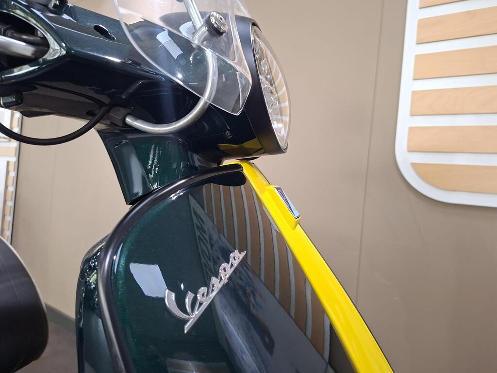 Vespa GTS 300 Racing Sixties 2022 3 Close-up of a vintage Vespa scooter with a glossy green body and yellow accents, featuring a front headlight and clear windshield, set against a neutral background.